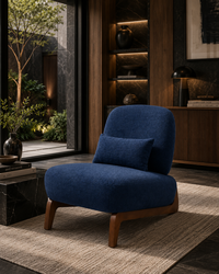 A modern blue upholstered accent chair with a matching pillow and wooden legs, styled in a contemporary, dimly lit living room setting.