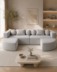 A light gray modular sectional sofa with circular back cushions and two ottomans, styled in a minimalist beige living room with a low wooden coffee table and built-in shelving.
