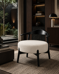 A modern lounge chair with a black curved wooden frame and a round off-white upholstered seat, placed in a dark, stylishly decorated room with a window view of a garden.