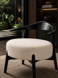 A modern accent chair with a thick, round beige upholstered seat cushion and a sleek, curved black wooden frame and legs, set on a textured rug in a dark, stylishly decorated interior.