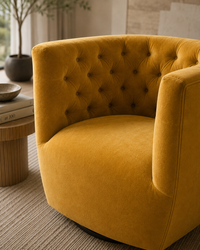 A mustard yellow velvet armchair with a tufted button-back design, shown in a modern, softly-lit interior setting next to a wooden side table.