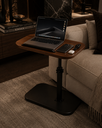 A black adjustable standing laptop table with a wooden top, shown beside a beige sofa in a modern living room. A laptop, phone, and earbud case are placed on the table.