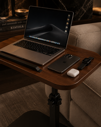 A wooden laptop table with a black metal leg base, styled in a cozy living room setting next to a sofa. On the table, there is an open laptop, a smartphone, a smartwatch, a white earbud case, and a slim black accessory.