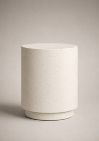 A minimalist cylindrical side table or lamp in a light beige speckled stone or concrete finish, set against a neutral warm gray background.