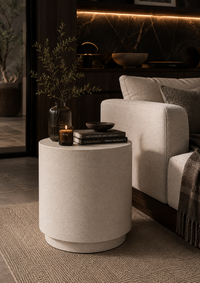A cylindrical side table in a light beige textured finish, styled in a modern living room next to a light gray sofa. The table is decorated with a dark glass vase containing dried branches, a lit candle, a small black bowl, and a stack of books.