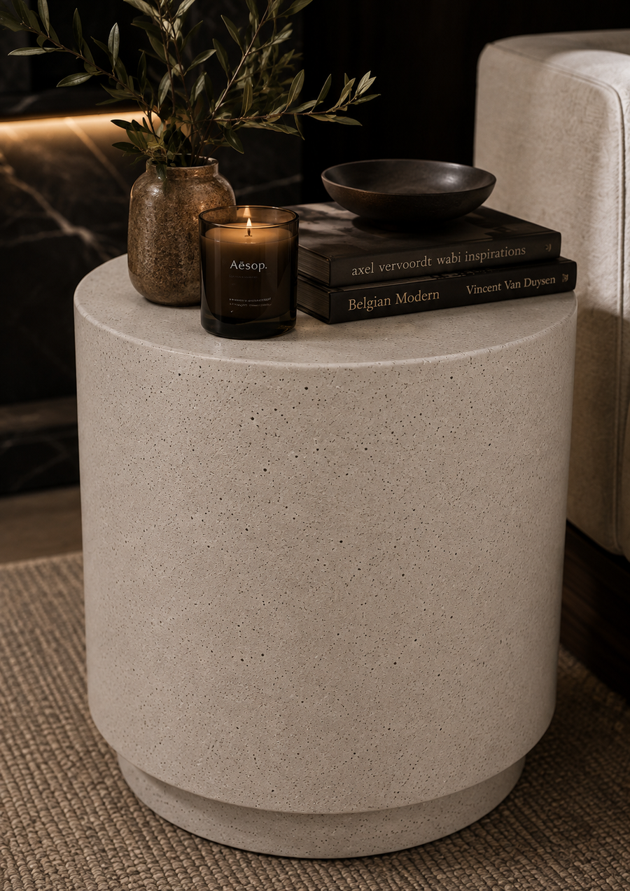 A cylindrical concrete side table in a warm light gray-beige tone, styled in a modern interior with a lit Arnoap candle, a small vase with greenery, books, and a bowl.
