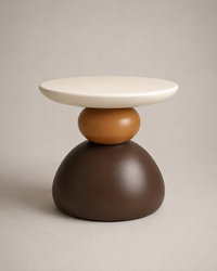 A modern sculptural side table with a wide, shallow oval top in off-white, a medium brown sphere in the middle, and a large dark brown dome-shaped base, set against a neutral warm gray background.