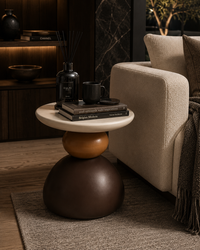 A modern round side table with a stacked sculptural design, featuring a white top, a terracotta middle sphere, and a dark brown base, styled in a cozy living room next to a beige sofa.