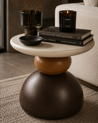A modern round side table with a stacked-bulb pedestal base in cream, terracotta, and dark brown, styled in a living room setting with books, a candle, and a small bowl.