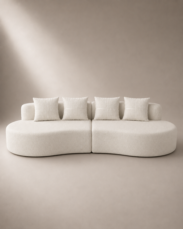 A modern curved modular sofa upholstered in a textured off-white fabric, featuring four matching square throw pillows, set against a minimalist beige background with soft diagonal lighting.