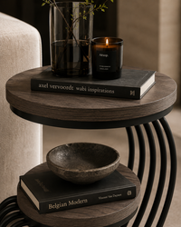 A round two-tier wooden side table with black metal curved legs, styled with a dark book, a glass bowl, a lit amber candle, and a tall glass vase in a cozy interior setting.