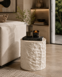 A cylindrical side table with a textured, rough-hewn stone finish in an off-white/cream color, styled in a modern living room next to a beige sofa. On top of the table are a black book, a clear glass vase with greenery, and a black candle jar.