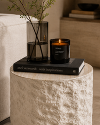 A lit black scented candle in a dark glass jar sits on a black book atop a textured white stone side table, with a tall smoky glass vase and green stems beside it in a softly lit interior setting.
