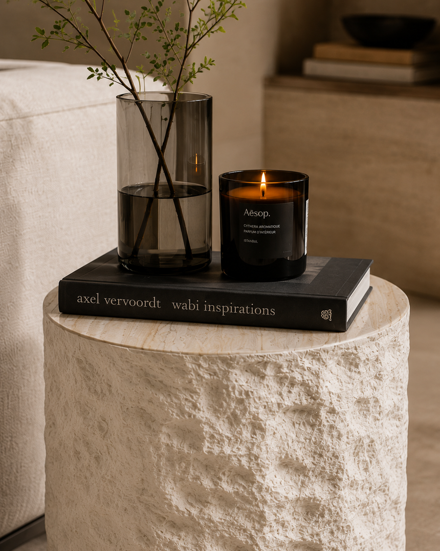 A lit black scented candle in a dark glass jar sits on a black book atop a textured white stone side table, with a tall smoky glass vase and green stems beside it in a softly lit interior setting.