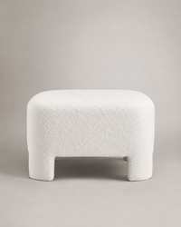 A modern low stool with a rounded rectangular seat and short legs, upholstered in a textured boucle fabric in off-white, set against a neutral light gray background.
