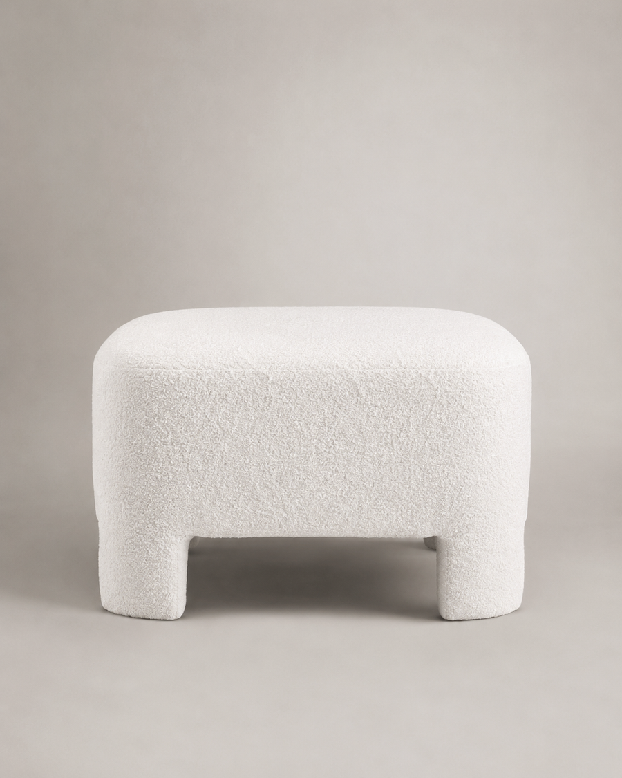A modern low stool with a rounded rectangular seat and short legs, upholstered in a textured boucle fabric in off-white, set against a neutral light gray background.