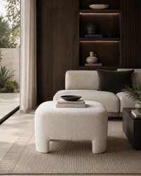 A modern living room scene featuring a textured off-white boucle ottoman in the foreground, with a matching light beige sectional sofa and dark charcoal accent pillow in the background. The room has dark wood shelving with warm ambient lighting, a woven beige rug, and a glass sliding door on the left.