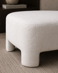 A close-up of a modern upholstered ottoman or stool with a textured bouclé fabric in off-white, shown on a neutral carpet in an indoor setting.