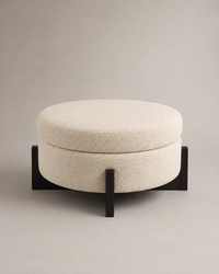 A round upholstered ottoman stool with a textured cream bouclé cushion and a black metal base, photographed against a neutral warm gray studio background.