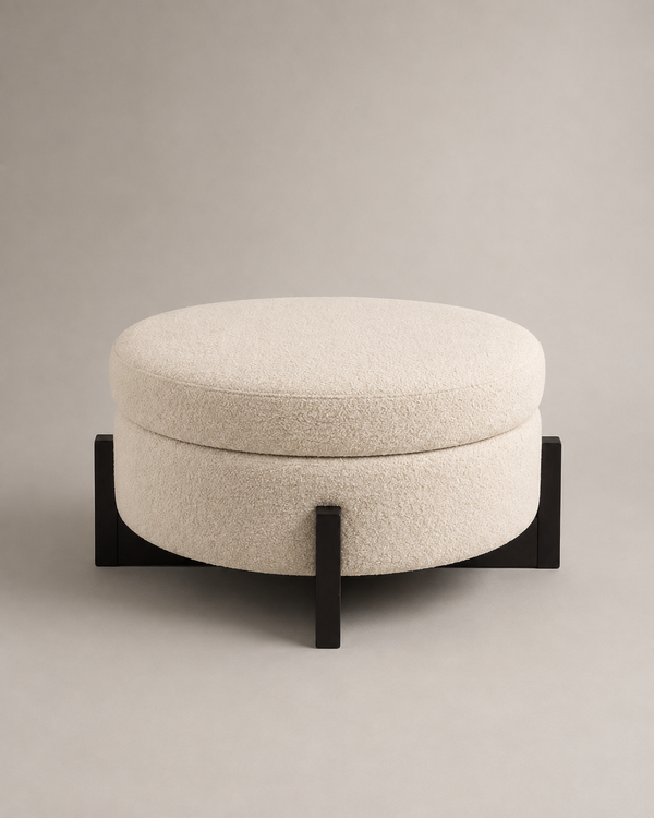 A round upholstered ottoman stool with a textured cream bouclé cushion and a black metal base, photographed against a neutral warm gray studio background.