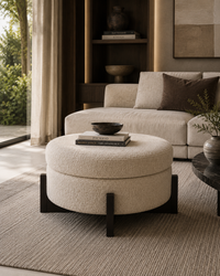 A round upholstered ottoman with dark wooden legs stands in a styled living room in front of a cream sofa. The ottoman has a textured bouclé-like surface and is styled with a small bowl and books on top.