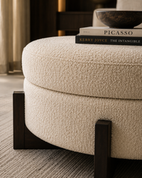 A round upholstered ottoman in textured cream bouclé fabric with dark wood block legs, shown in a styled interior setting with books and a bowl on top.