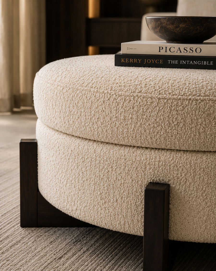 A round upholstered ottoman in textured cream bouclé fabric with dark wood block legs, shown in a styled interior setting with books and a bowl on top.