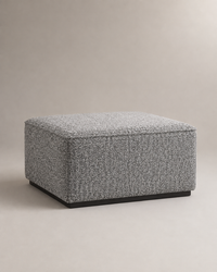 A square upholstered ottoman in a textured gray and white woven fabric, featuring a black base, set against a neutral beige background.