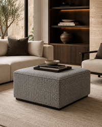 A square upholstered ottoman in textured gray fabric sits in a modern living room setting, styled with a stack of books and a small bowl on top. The room features a cream sofa, a dark wood shelving unit with warm lighting, and a beige rug.