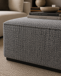 A square upholstered ottoman in a textured gray and white heather fabric, shown in a styled living room setting with books and a bowl on top.