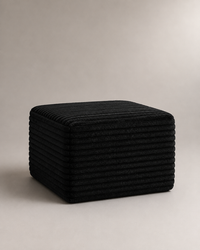 A black corduroy cube ottoman or stool with a ribbed fabric texture, photographed on a neutral light gray studio background.