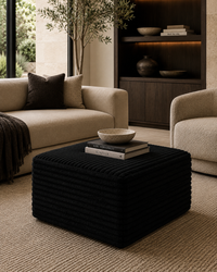 A black ribbed cube ottoman in a modern living room setting, styled with a book and bowl on top, positioned between a beige sofa and an armchair.