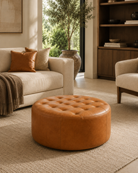 A round tufted leather ottoman in a warm cognac brown sits on a textured beige rug in a modern, sunlit living room with a cream sofa and wood shelving.