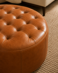 A close-up of a round tufted leather ottoman in a warm cognac brown, positioned on a textured beige rug with part of a sofa leg visible in the background.