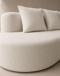 A close-up of a modern curved sofa upholstered in textured cream bouclé fabric, with two matching square throw pillows against a neutral beige wall.