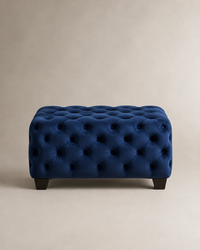 A rectangular tufted ottoman in deep navy blue velvet, supported by short dark wooden legs, set against a neutral beige background.
