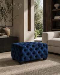 A deep blue tufted velvet ottoman with short dark legs, styled in a modern living room setting next to a beige sofa and a dark wood cabinet.