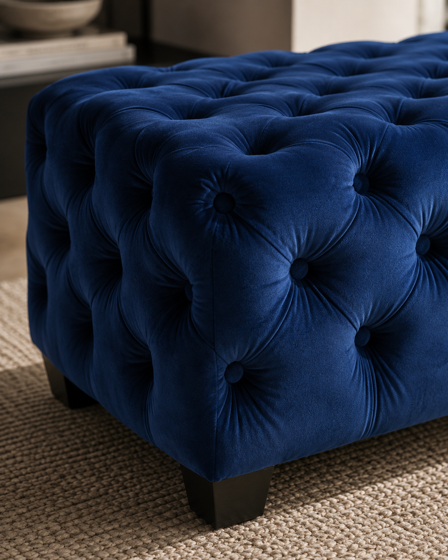 A close-up of a deep blue tufted velvet ottoman with dark wooden legs, resting on a textured beige woven rug.