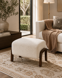 A modern boucle ottoman with dark wood legs in a styled living room setting next to a white sofa with a brown throw blanket.