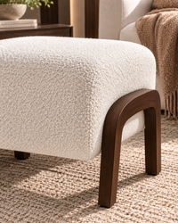 A close-up of a modern upholstered ottoman with a textured cream bouclé seat and dark brown wooden legs, set on a woven rug in a styled living room.