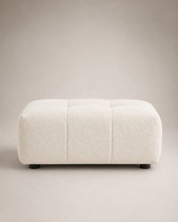 A modern low rectangular ottoman upholstered in a textured bouclé fabric, shown in a warm off-white/cream color. It features tufted panel detailing on the top and small dark feet at the bottom, set against a neutral beige studio background.