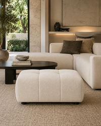 A modern living room scene featuring a cream bouclé ottoman in the foreground, a matching cream sectional sofa with dark brown and beige pillows, a dark wood coffee table, and a textured beige rug. A large window on the left reveals greenery outside.