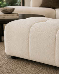 A close-up of a cream bouclé ottoman in a modern living room setting, showing its soft textured fabric and tufted panel design.
