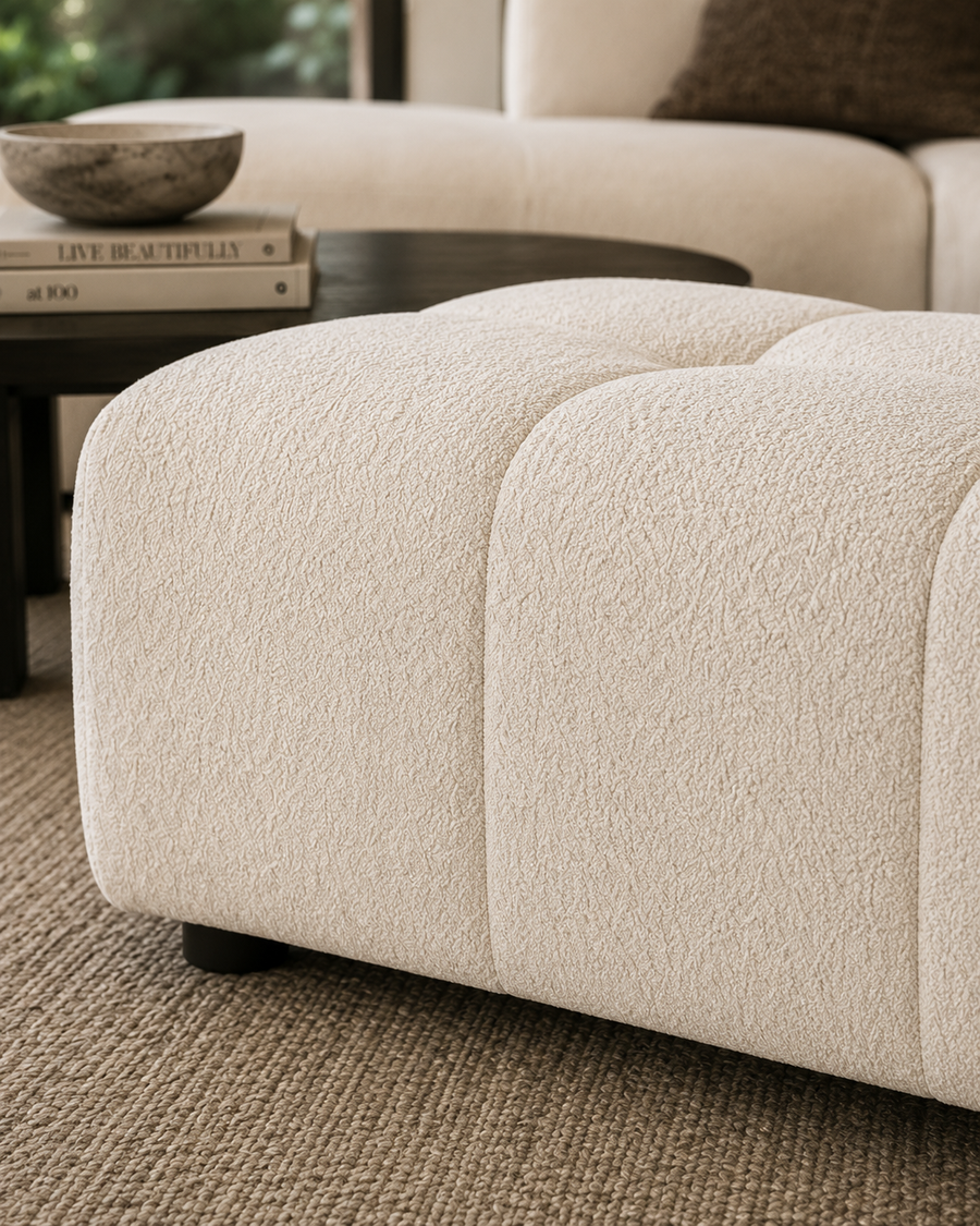 A close-up of a cream bouclé ottoman in a modern living room setting, showing its soft textured fabric and tufted panel design.