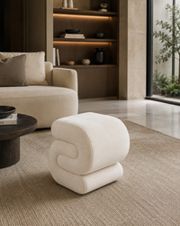 A modern, sculptural off-white upholstered ottoman with a stacked, wavy design, placed on a textured beige rug in a contemporary living room setting.