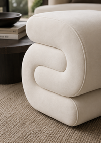 A close-up of a cream-colored upholstered ottoman with a sculptural, wavy channel design, set on a textured beige rug next to a dark wooden side table.