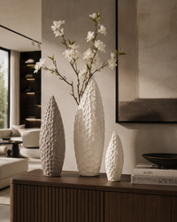 Three textured white ceramic vases of different heights arranged on a dark wood console table in a modern living room setting, with white blossoms in the tallest vase.