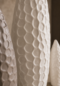 A close-up of a tall, textured ceramic vase with a raised honeycomb pattern, shown alongside other matching vases in a warm, neutral setting.