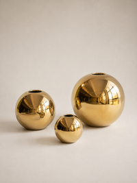 Three reflective gold metallic spherical vases of different sizes arranged on a light beige studio background.