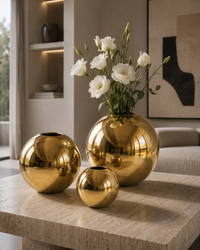 Three shiny gold spherical vases arranged on a stone table in a modern living room, with white flowers placed in the tallest vase.